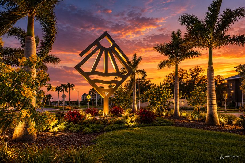 Lit Wellen Park logo sculpture, a sun over waves, among palms and tropical plantings at dusk